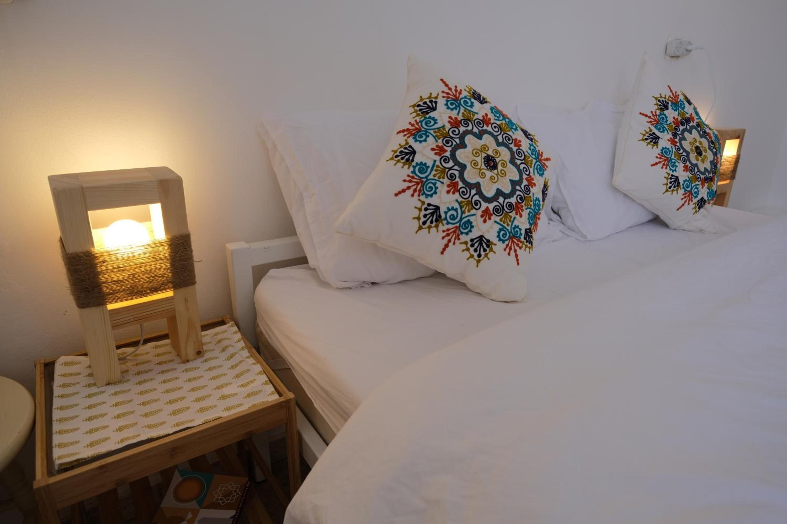 Una cama bien hecha con sábanas blancas y almohadas decorativas con diseños intrincados. Una pequeña mesita de noche con lámpara está colocada al lado de la cama.