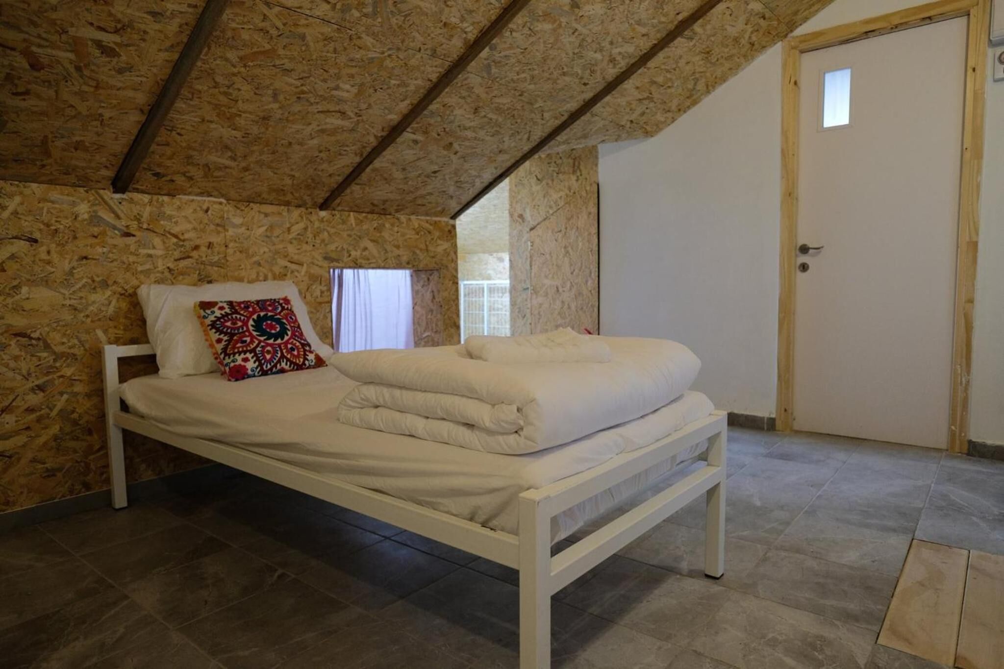 A bedroom with a wooden ceiling, a bed with white bedding, a colorful pillow, and a door.