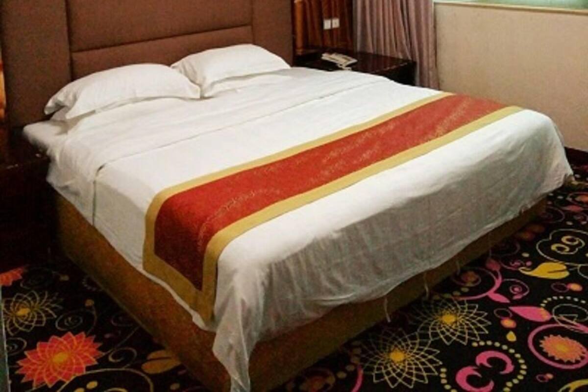 A hotel room with a large bed, a headboard, a bedside table, a lamp, a window with curtains, and a carpet with a floral pattern.
