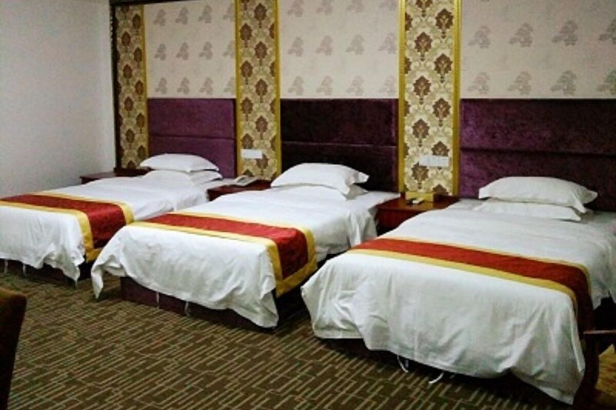 A hotel room with four single beds, each with a white bedspread and red and yellow trim. The room has a patterned carpet and a large, ornate headboard.