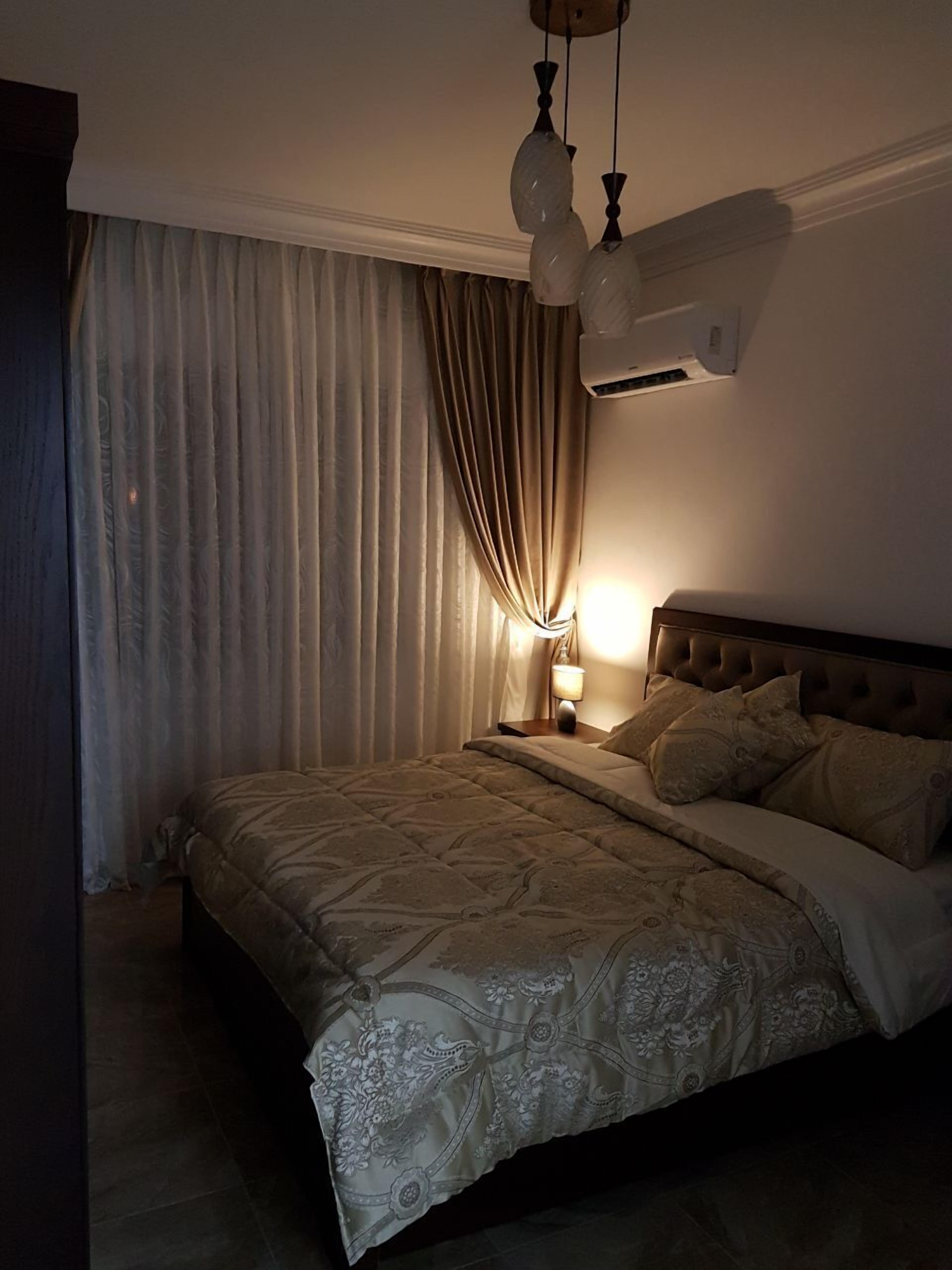 Un dormitorio con cama, cortinas, mesita de noche, lámpara y aire acondicionado.