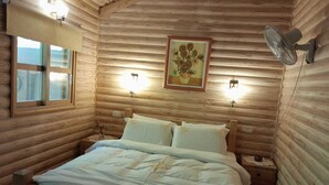 Une chambre dans un chalet en bois, avec un lit, deux tables de chevet, un ventilateur fixé au mur et un tableau encadré au-dessus du lit.