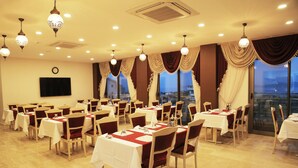 Un salón de banquetes con varias mesas dispuestas para comer, ventanas amplias con cortinas y candelabros colgantes.