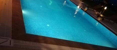 Una piscina nocturna con sillones de descanso y sombrillas alrededor.