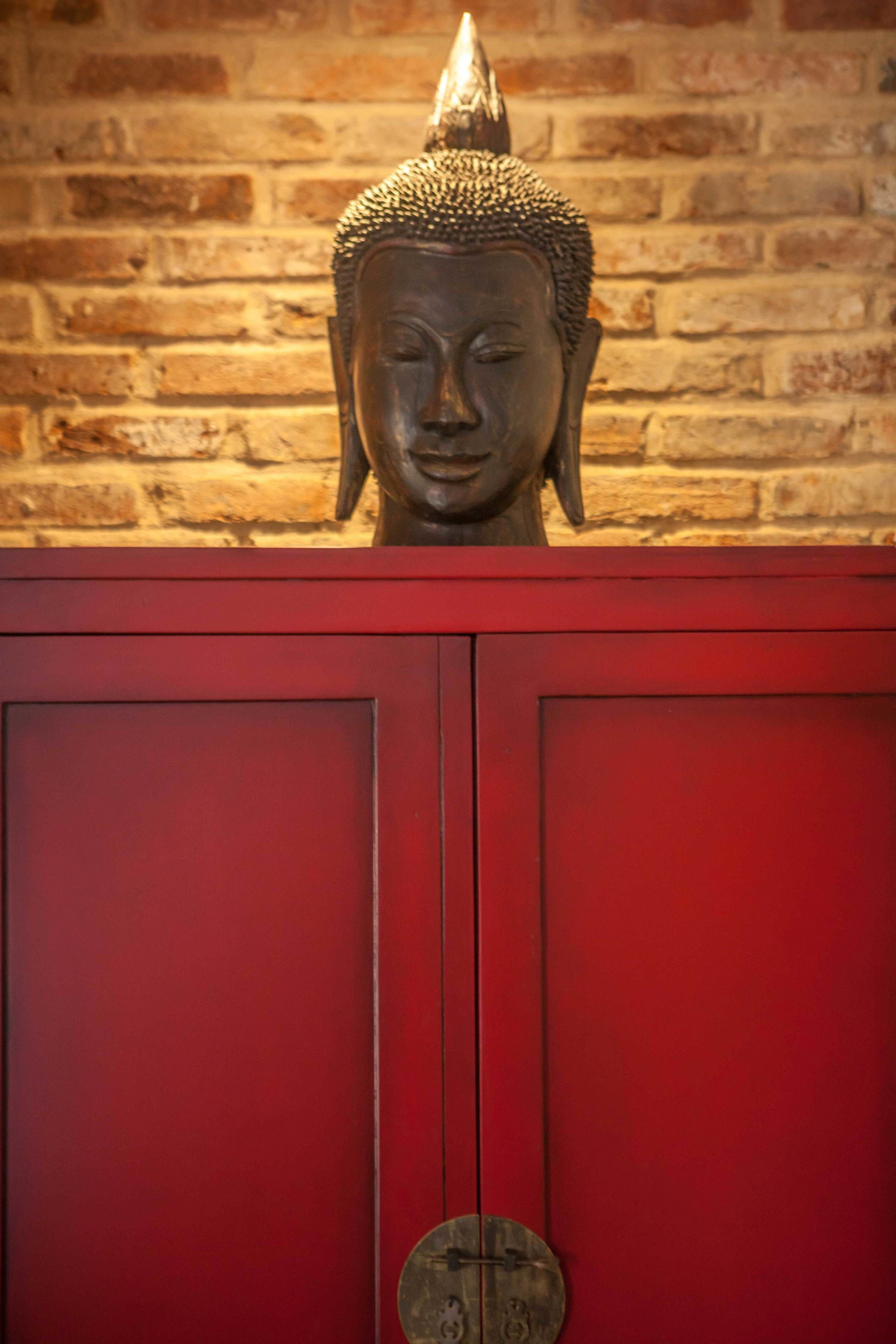 Une statue de Bouddha, placée sur un meuble rouge, se dresse devant un mur de briques.