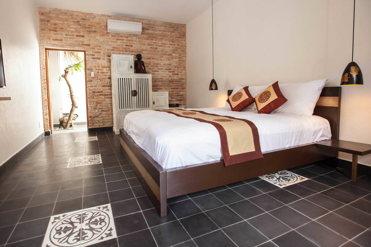 A hotel room with a brick accent wall, a large bed with white bedding and decorative pillows, a white wardrobe, and a television mounted on the wall.