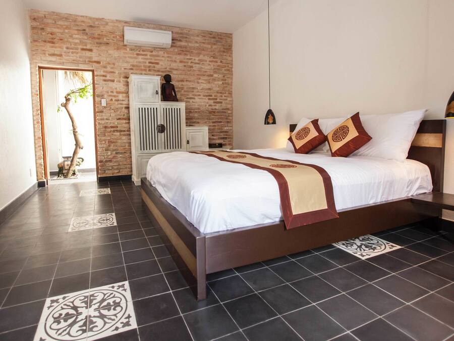 A hotel room with a brick accent wall, a large bed with white bedding and decorative pillows, a white wardrobe, and a television mounted on the wall.