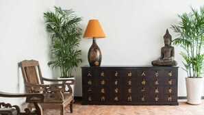 A room with a wooden bench, a dark wooden cabinet with drawers, a lamp, and two potted plants. - La Casita Saigon (Ho Chi Minh City)