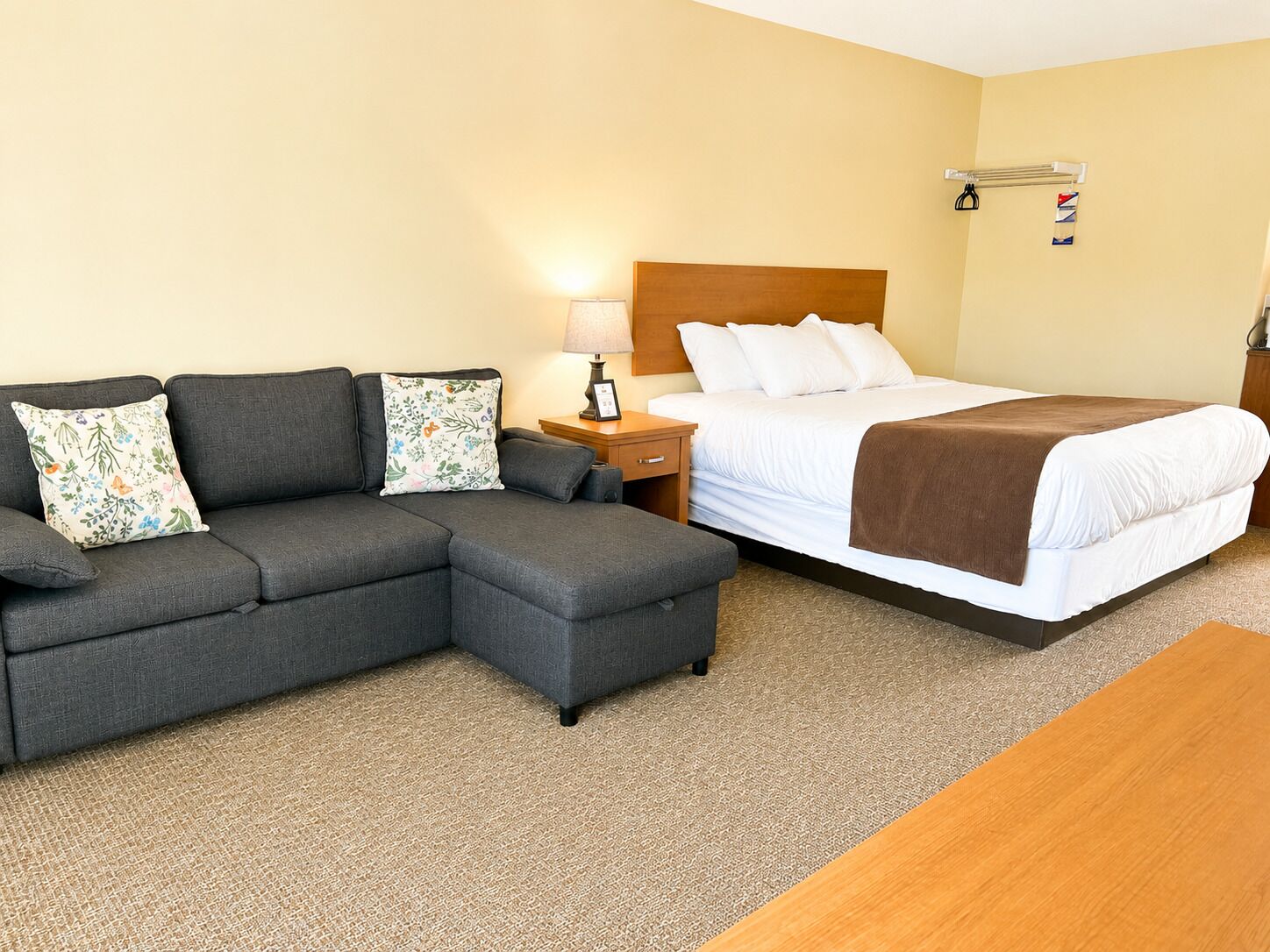 A hotel room with a sectional sofa, a single bed, a wooden nightstand, a lamp, and a small table.