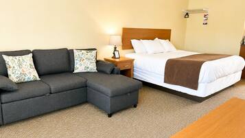 A hotel room with a sectional sofa, a single bed, a wooden nightstand, a lamp, and a small table.