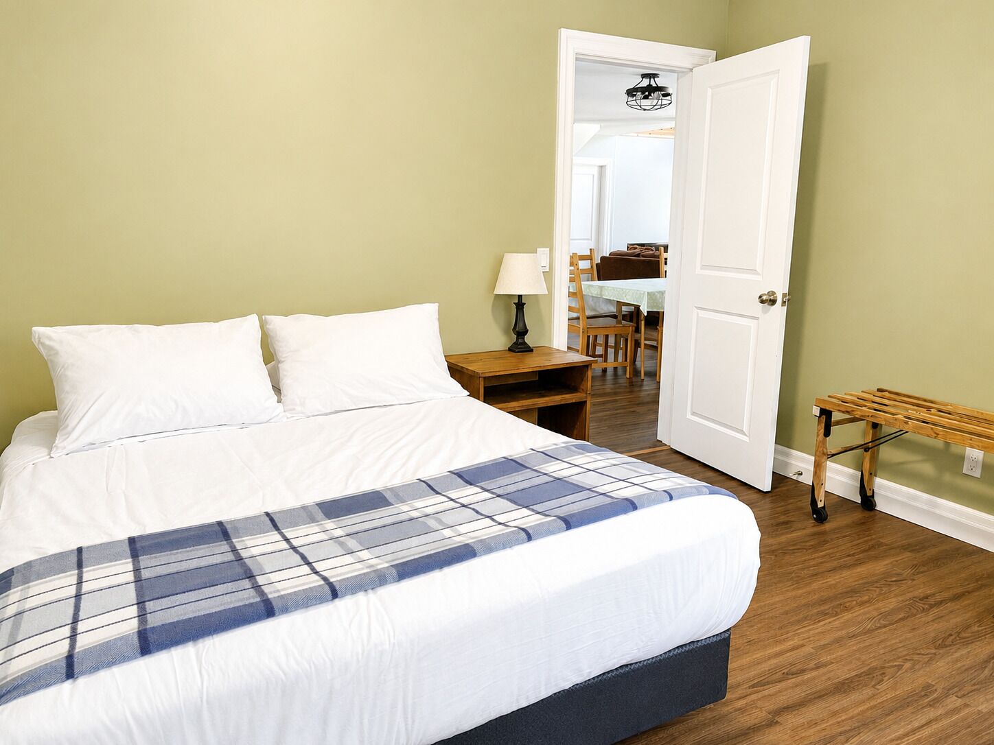 A bedroom with a bed, a nightstand, a lamp, and a door leading to a dining area.