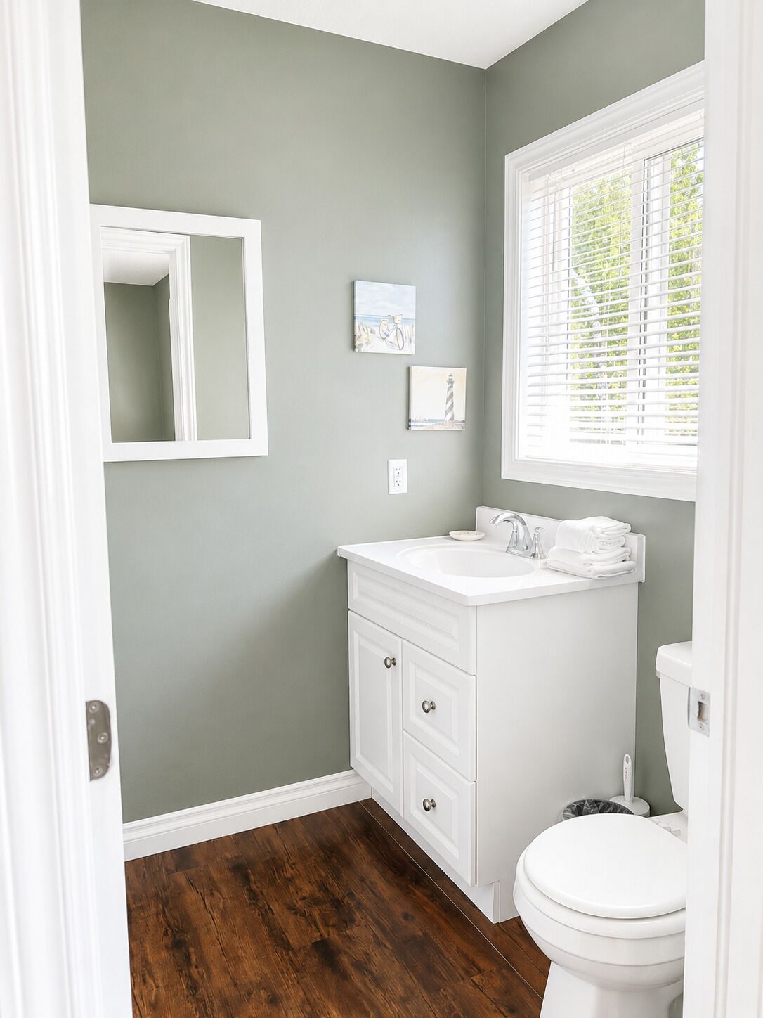 A bathroom with a white vanity, a toilet, a window with blinds, and two wall-mounted artworks.