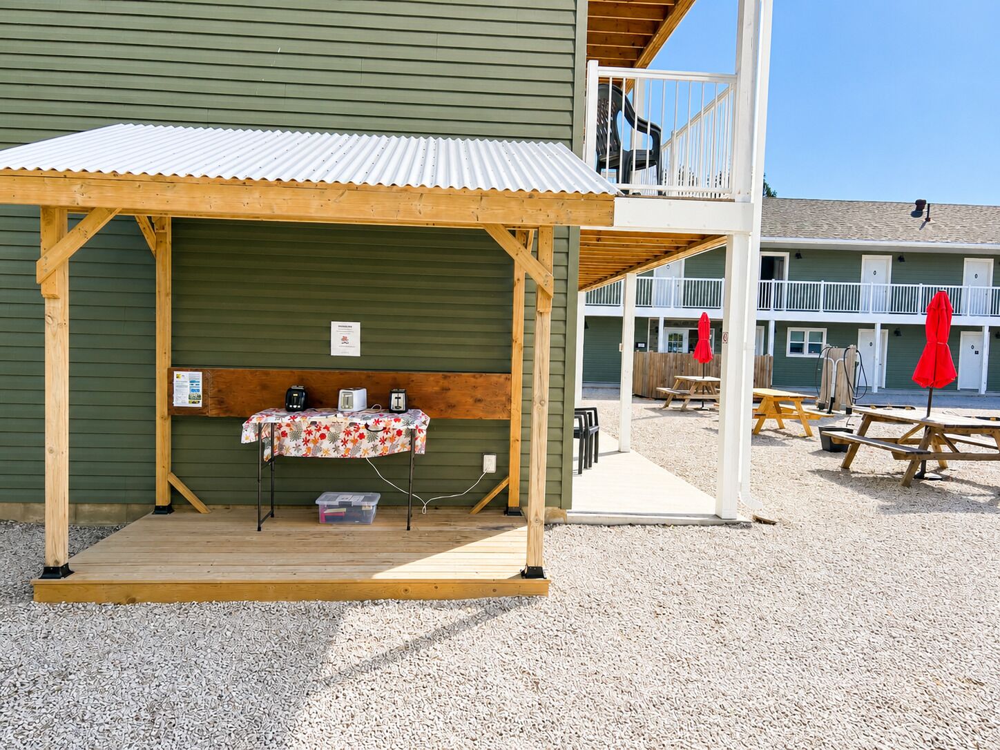 Une structure en bois comprenant une table et des prises électriques, un espace abrité et, au fond, un bâtiment doté d’un balcon.