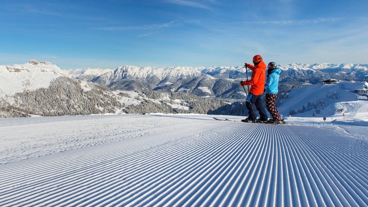 A snowy ski slope with two skiers in the foreground and a mountain range in the background.