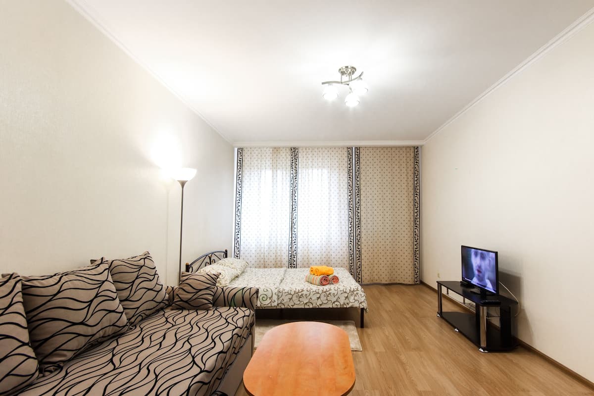 Una habitación pequeña y luminosa con una cama, un sofá, un televisor sobre un mueble y una lámpara.