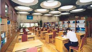L’intérieur d’une bibliothèque, avec de nombreuses tables et chaises, des étagères et un plafond orné de luminaires circulaires.