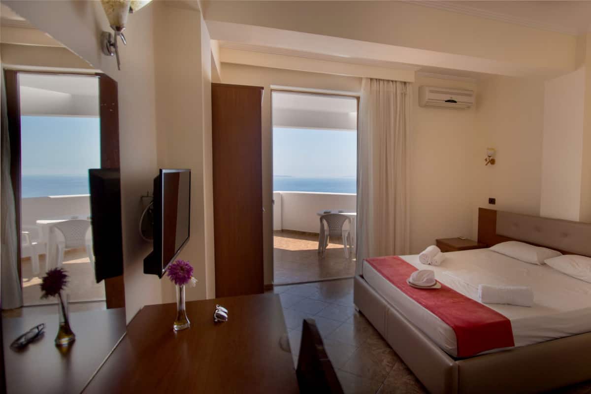 Een hotelkamer met een bed, een televisie, een bureau met een vaas en een balkon met uitzicht op zee.