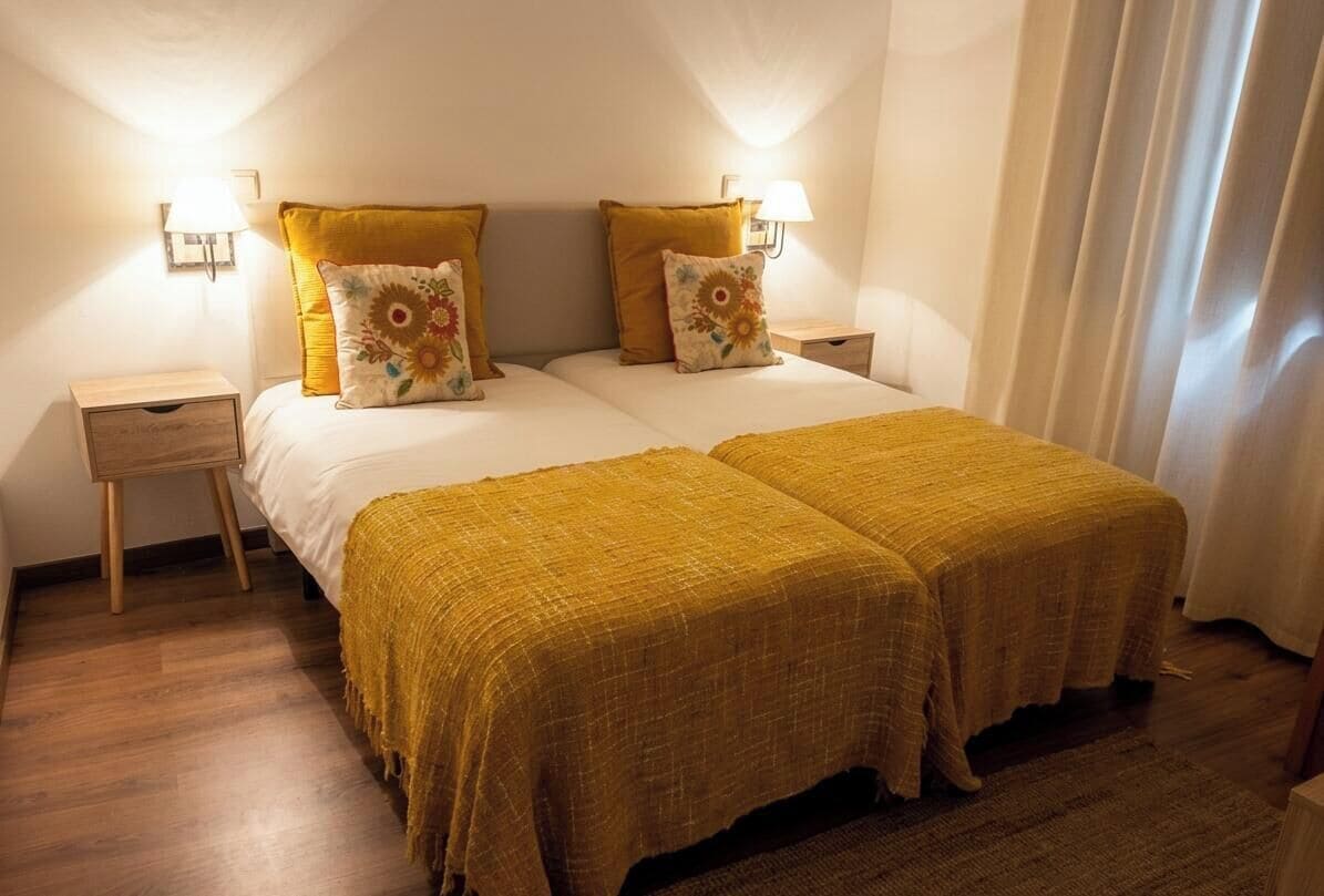A double bed with a yellow bedspread and two pillows, a nightstand with a lamp, and a window with curtains.