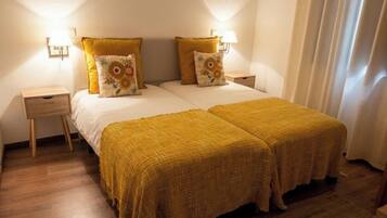 A double bed with a yellow bedspread and two pillows, a nightstand with a lamp, and a window with curtains.