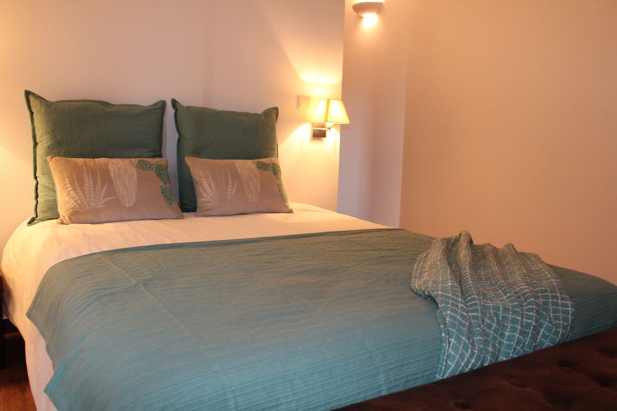 A neatly made bed with a teal bedspread and multiple pillows, a wall-mounted lamp, and a dark brown headboard.