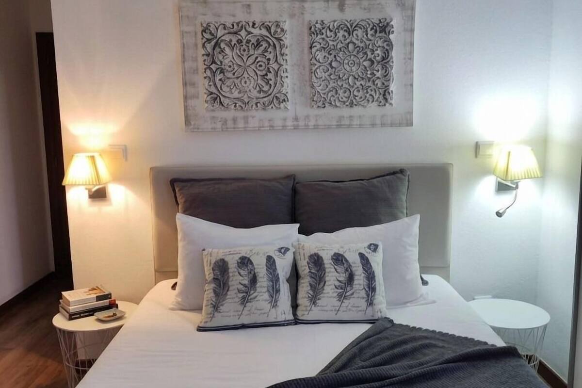 A bedroom with a bed, pillows, a bedside table, and wall-mounted lamps.