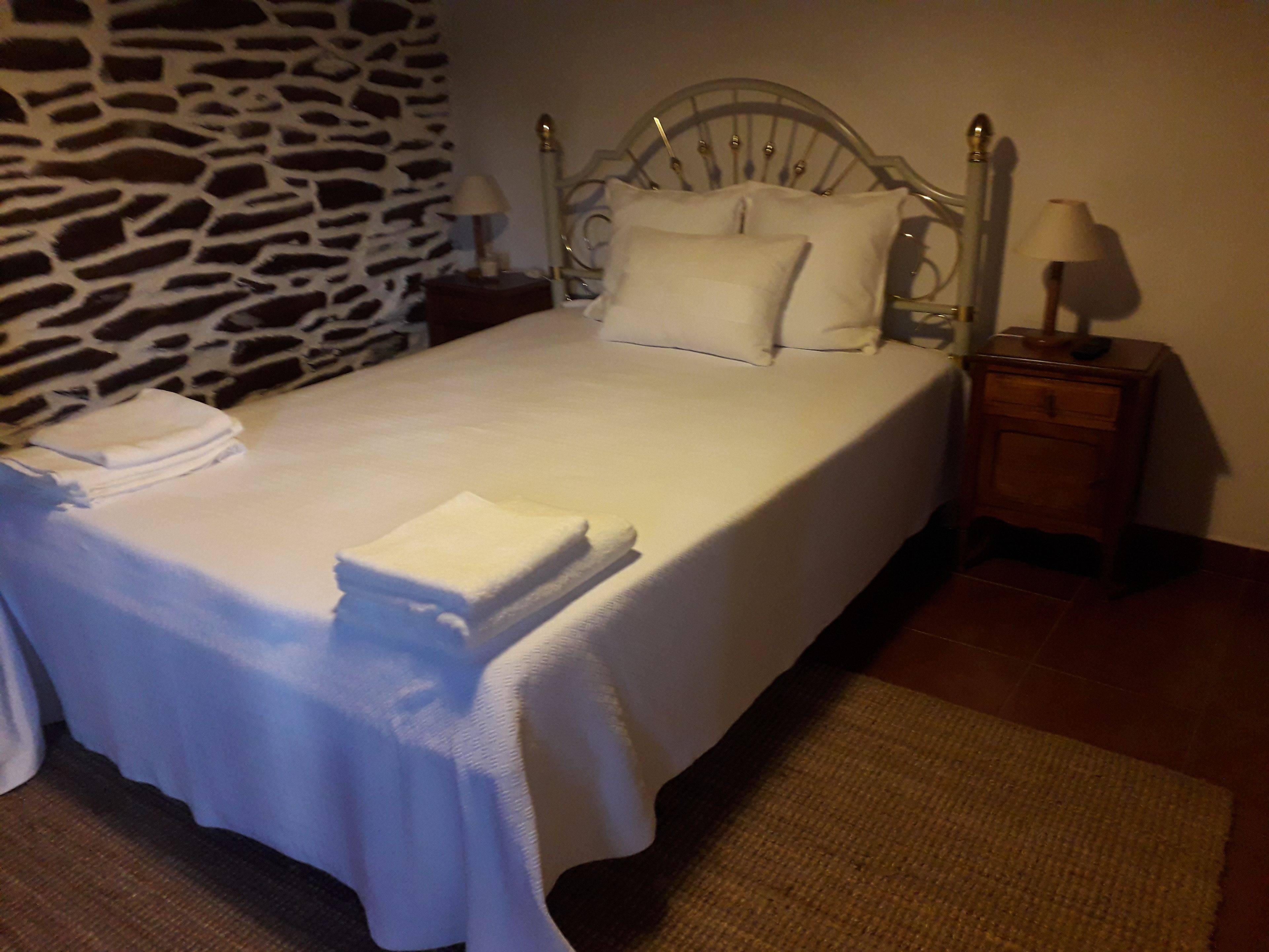 Een net opgemaakt bed met witte lakens, een muur met een stenen patroon en een klein houten nachtkastje met een lamp.