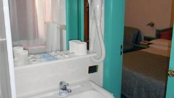 Baño con lavabo, espejo y toallero. Se ve una cama en la habitación contigua.