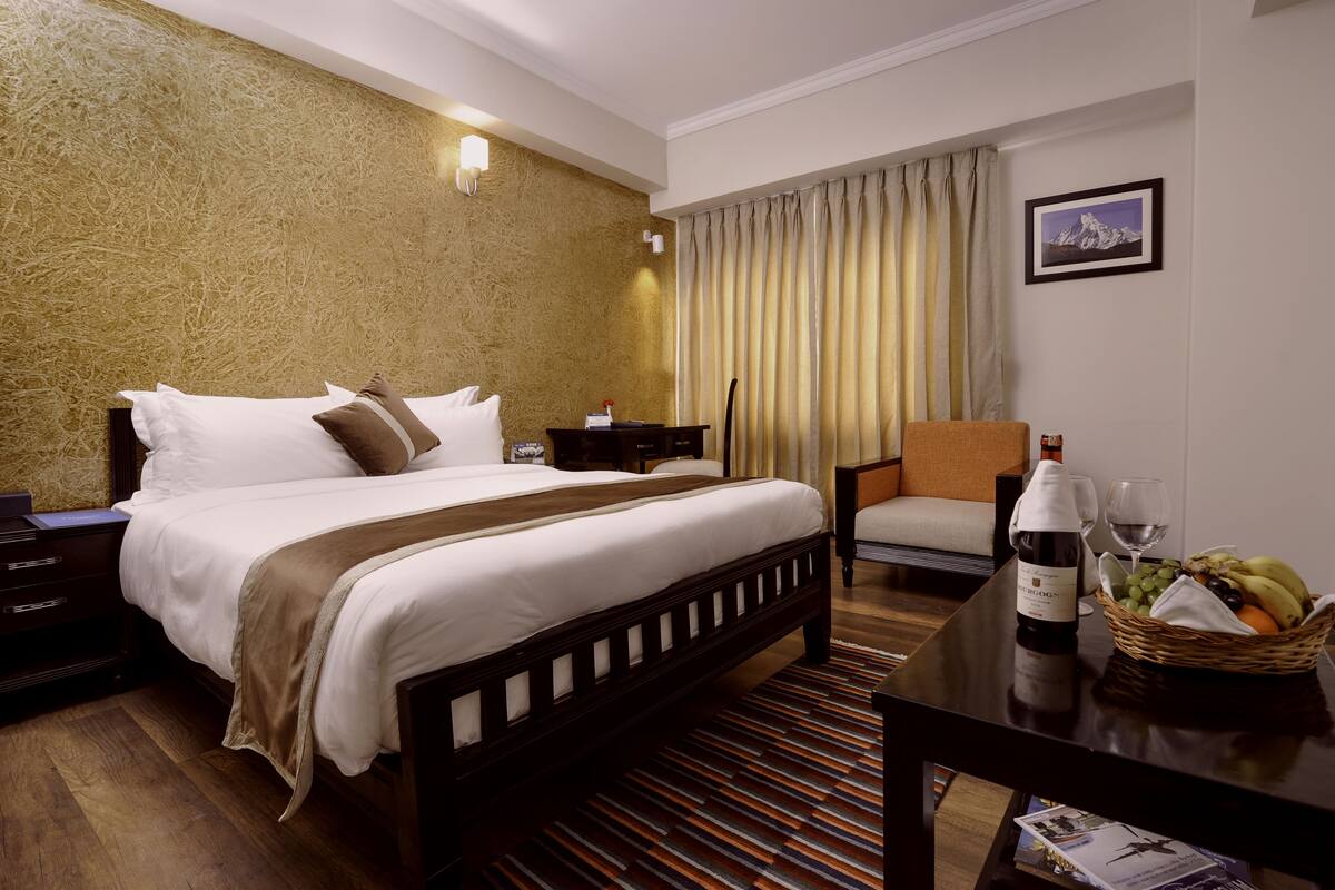 A hotel room with a large bed, a wooden nightstand, a chair, a table with a bottle and glasses, and a fruit basket.