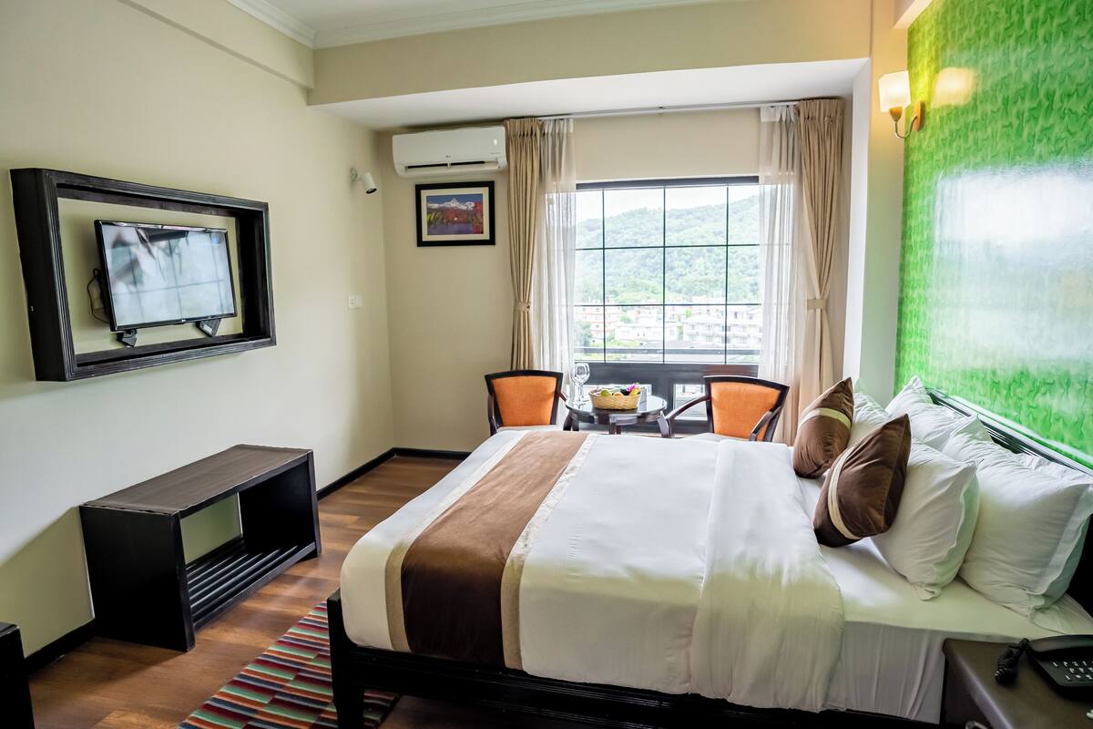 A hotel room with a bed, a TV, a small table with chairs, and a window with curtains.