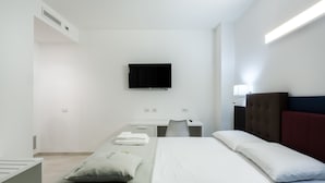 A modern hotel room with a bed, a desk, a chair, a flat-screen TV mounted on the wall, and a bedside table with a lamp. - TH Collection Rooms (Oristano)