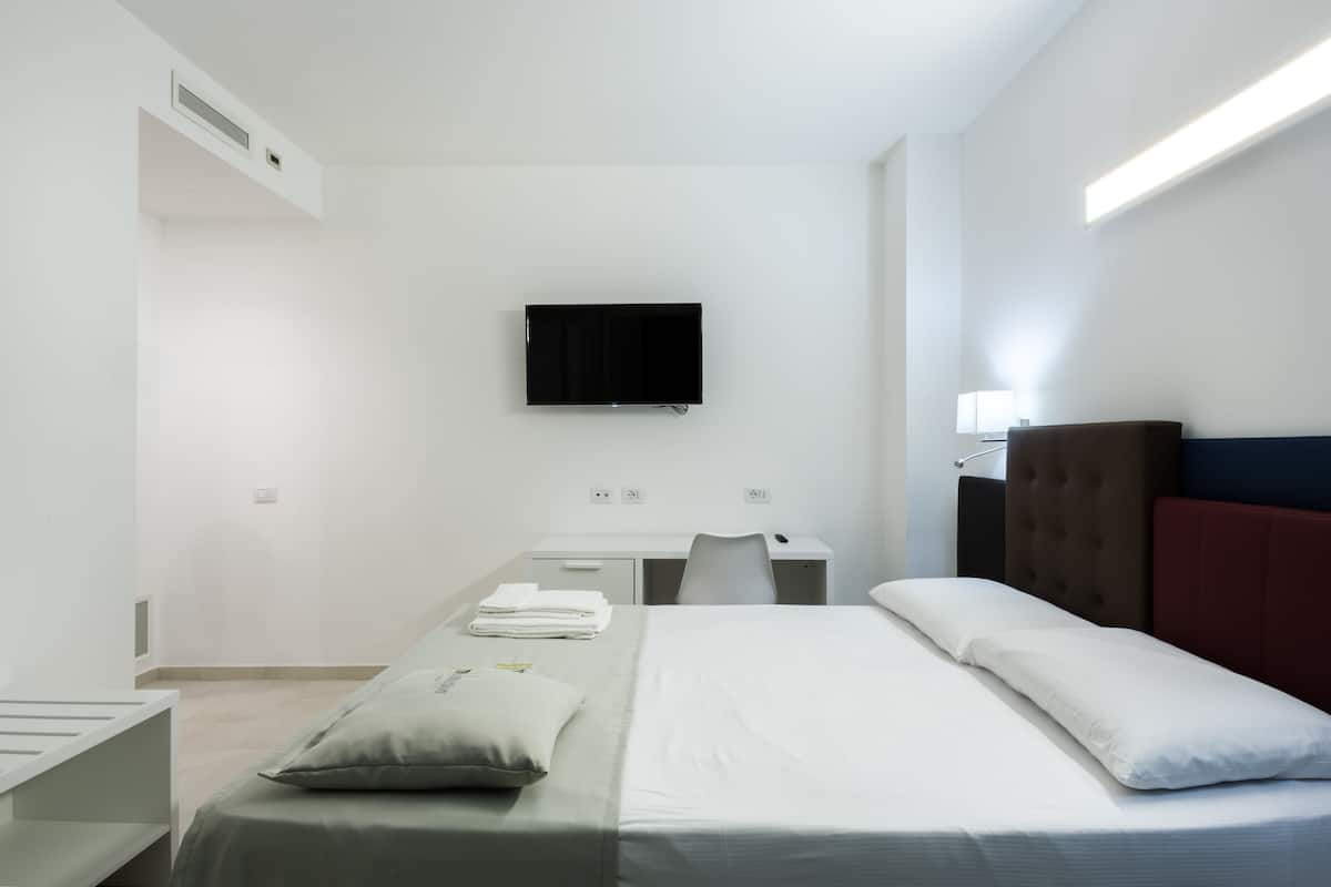 A modern hotel room with a bed, a desk, a chair, a flat-screen TV mounted on the wall, and a bedside table with a lamp.