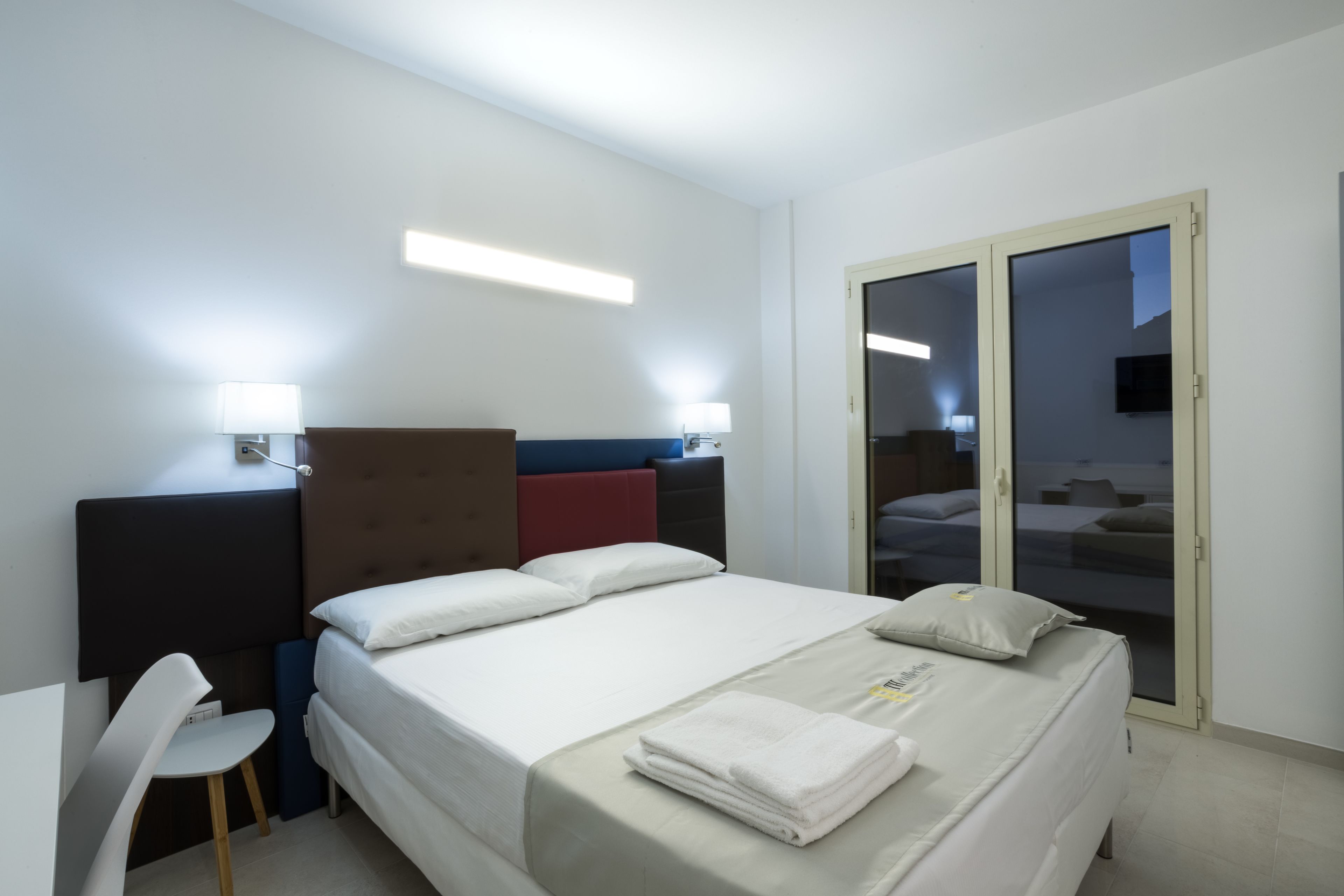 A hotel room with a bed, bedside tables, a chair, and a sliding glass door to a balcony.