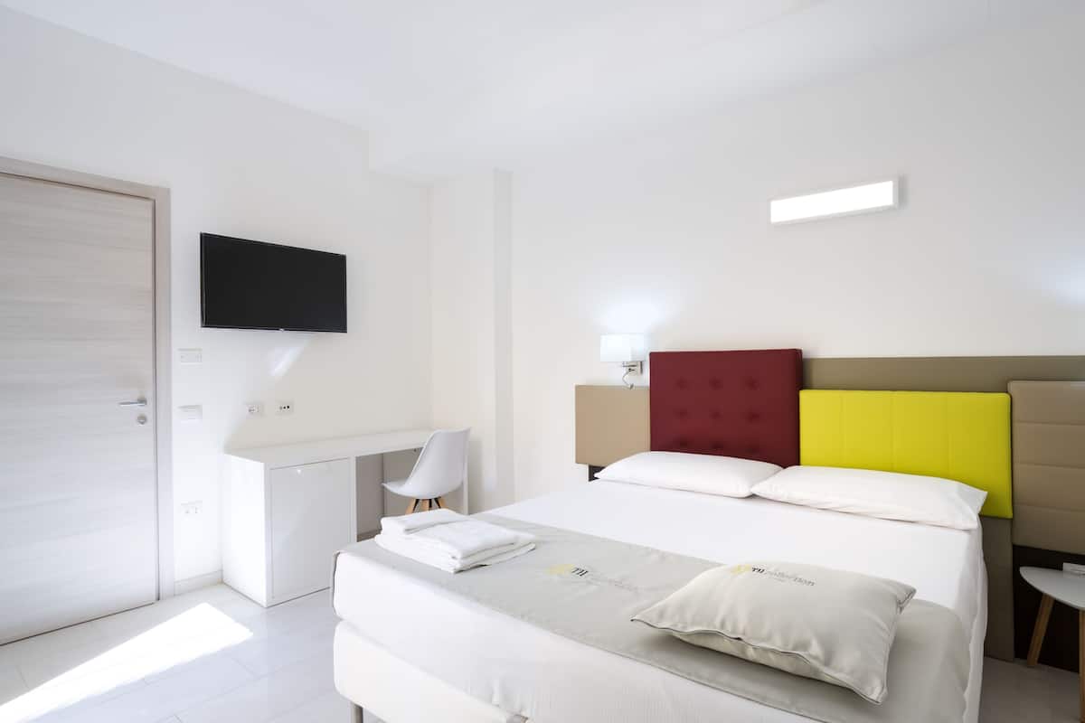 A modern hotel room with a bed, a desk, a chair, a TV, and a bedside table.