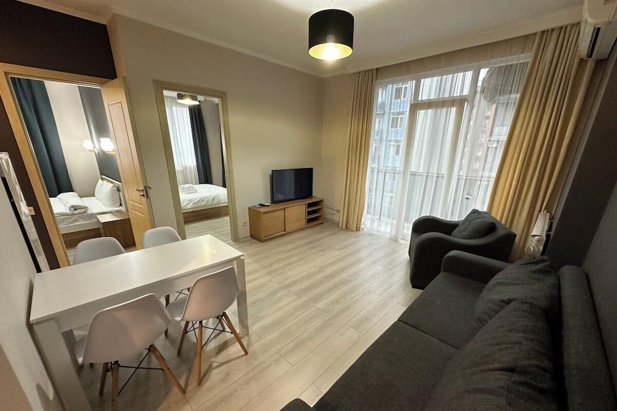 Una sala moderna con sofá, una mesa de comedor con sillas, televisor y cama al fondo.