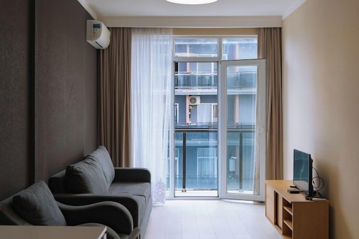 Una sala moderna con un sofá, una mesa de comedor y sillas, un televisor sobre un mueble bajo, y un amplio ventanal corredero que da a un balcón.
