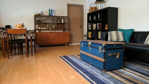 A living room with a wooden dining table, chairs, a blue suitcase, a black sofa, a striped rug, and a bookshelf.