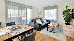 Een moderne woonkamer met een eettafel, een zwarte leren bank en een groot raam met dunne gordijnen.