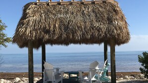 A beachside gazebo with a thatched roof, two white Adirondack chairs, and a small table. - Penthouse Apartment Sunsets in Key Largo, Kawama Yacht Club Tower (Key Largo)