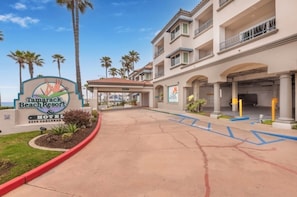 A hotel entrance with a sign, palm trees, and a parking area.
