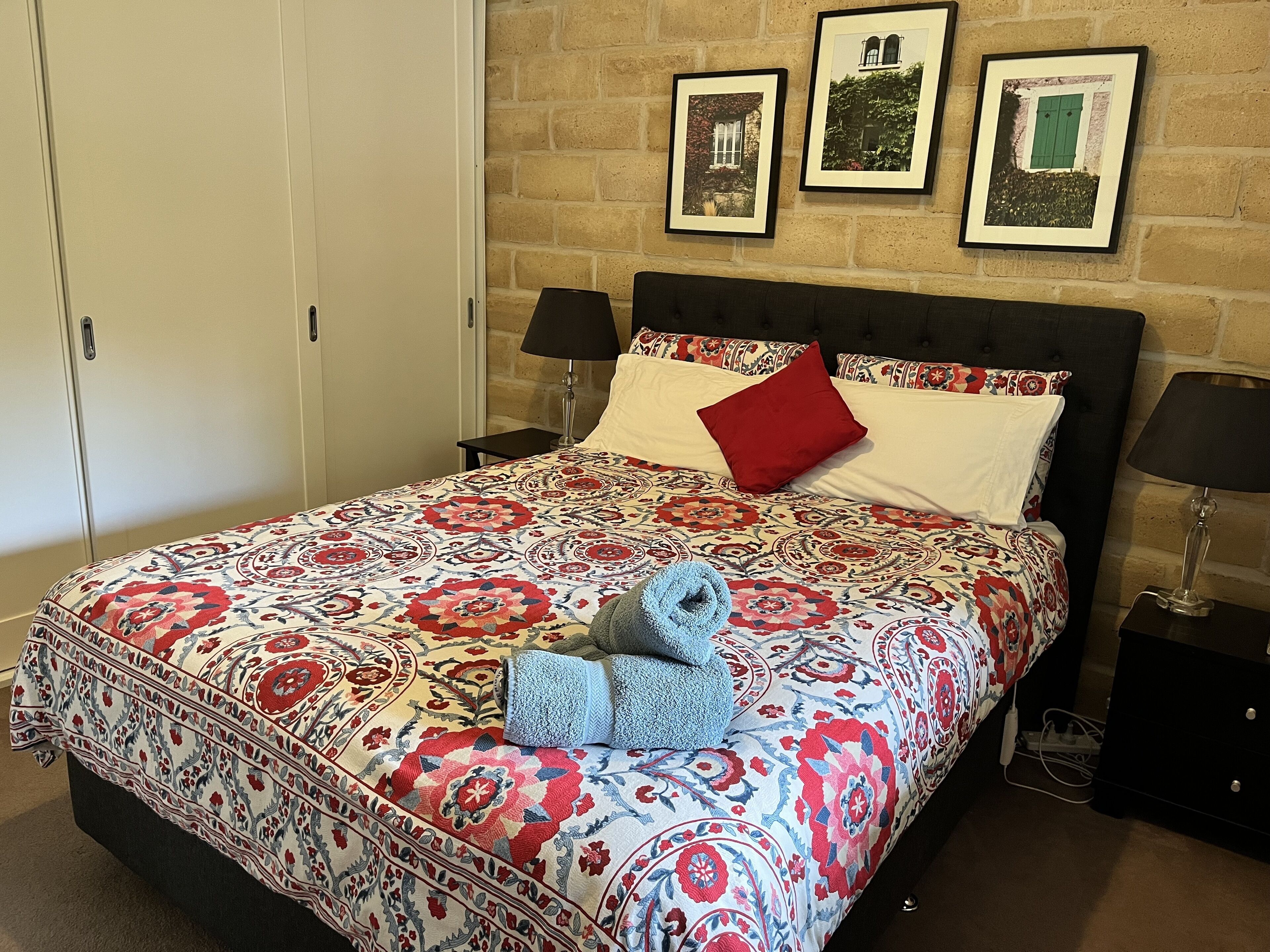 Un lit bien fait, recouvert d’une couvre-lit à motifs, avec deux lampes de chevet et un mur orné de trois tableaux encadrés.
