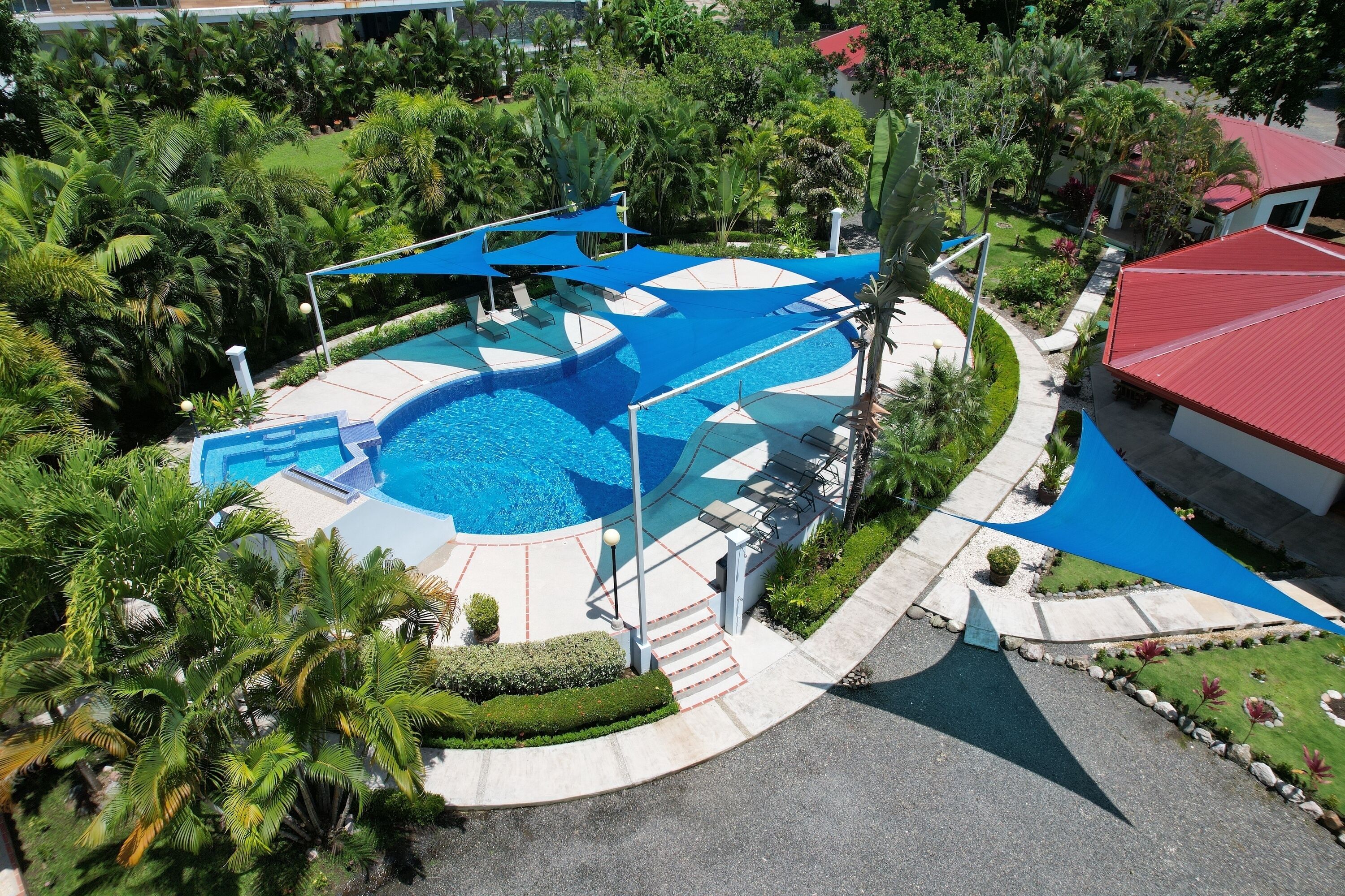 A swimming pool with a blue deck, surrounded by palm trees and a red-roofed building.