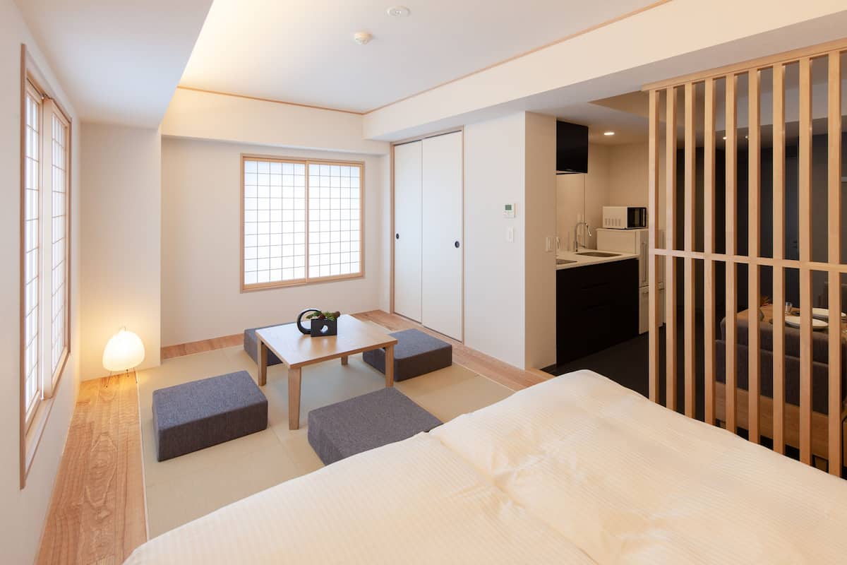 A modern hotel room with a bed, a small table, a bench, a window, and a kitchenette.