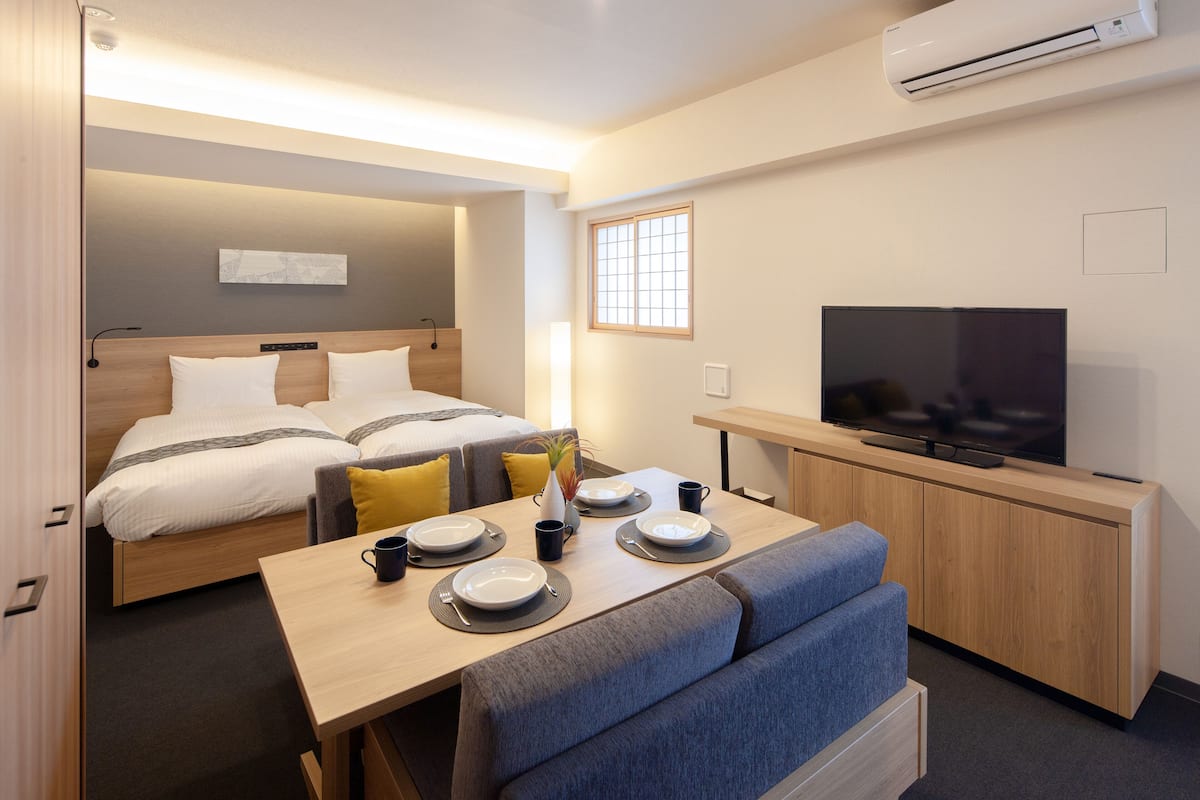 A modern hotel room with a bed, a dining table set with plates and cups, a sofa, a TV, and an air conditioning unit.