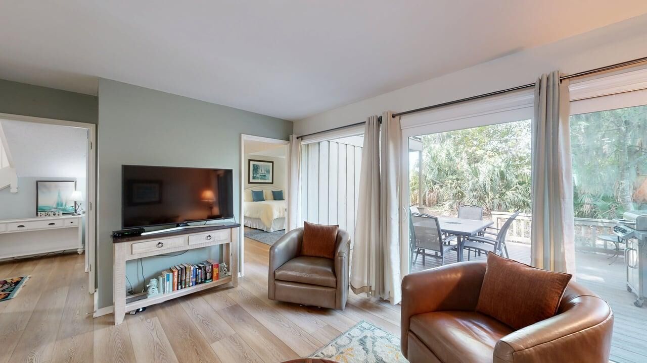 A modern living room with a flat-screen TV, leather armchairs, a bookshelf, and a sliding glass door leading to an outdoor patio.
