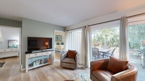 A modern living room with a flat-screen TV, leather armchairs, a bookshelf, and a sliding glass door leading to an outdoor patio. - Short walk or bike ride to the beach and Palmetto Dunes golf! (Hilton Head)
