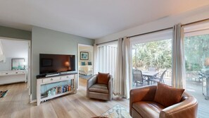A modern living room with a flat-screen TV, leather armchairs, a bookshelf, and a sliding glass door leading to an outdoor patio. - Short walk or bike ride to the beach and Palmetto Dunes golf! (Hilton Head)