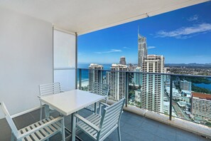 Un balcón con una mesa y sillas blancas, con vistas a un paisaje urbano con edificios altos y una masa de agua.