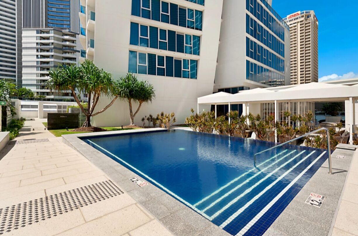 Une piscine à l’eau d’un bleu cristallin, entourée d’une zone pavée agrémentée de palmiers et d’un bâtiment blanc doté de grandes fenêtres.