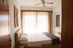 A bedroom with a bed, a ceiling fan, a window with curtains, a bedside table, and a wall-mounted TV.