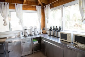A modern kitchen with stainless steel appliances, including multiple coffee makers, a microwave, and a toaster. There are large windows with curtains, providing natural light and a view of greenery outside.
